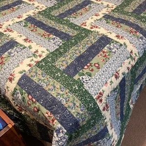 May include: A quilted bedspread with a patchwork design. The quilt features alternating stripes of floral patterns in cream, blue, and burgundy, and solid navy and green. The quilt is made of a soft, quilted fabric.