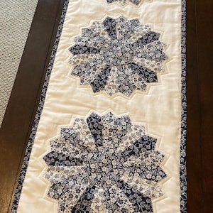 May include: A white and navy blue floral patterned table runner. The runner features three large, circular floral designs in navy blue and white, set against a white background. The edges are trimmed with a navy blue floral border.
