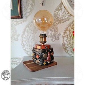 Könnte beinhalten: Eine Tischlampe mit einer großen, runden, bernsteinfarbenen Edison-Glühbirne auf einem dunklen, quadratischen Sockel. Der Sockel ist mit verschiedenen Metallzahnrädern, einer kleinen Fuchskonstruktion und dem Buchstaben W verziert, auf einem Holzbrett.