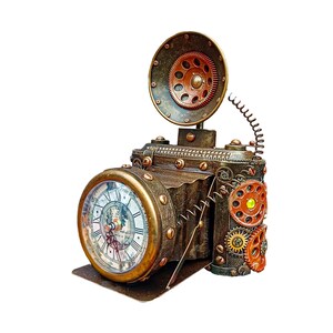 Puede incluir: Un reloj con forma de cámara de estilo steampunk, con acabado en bronce. El reloj presenta una esfera redonda con números romanos y un flash decorativo. Engranajes y remaches adornan el cuerpo de la cámara.
