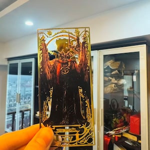 Op de afbeelding: Een goudkleurige tarotkaart met een illustratie van een duivelachtige figuur met hoorns en een lange jurk. De kaart is omlijst met sierlijk gouden filigraan en de tekst "THE DEVIL XV" is zichtbaar aan de onderkant.