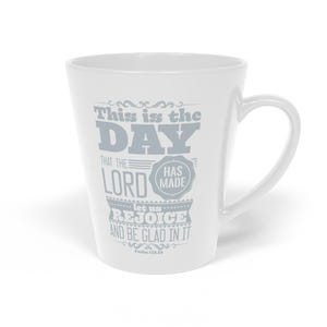 May include: A white ceramic latte mug featuring grey text that reads: This is the DAY that the LORD HAS MADE let us REJOICE AND BE GLAD IN IT Psalm 118:24. The mug has a tapered shape with a curved handle on the right side and a smooth finish.