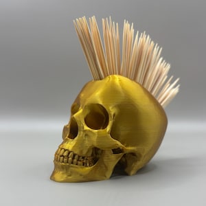 Puede incluir: Un porta palillos de dientes en forma de calavera de color dorado con un mohicano de palillos.
