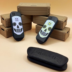 Puede incluir: Monopatines de dedo en blanco y negro con diseños de calaveras de azúcar. Los monopatines tienen una base negra y un gráfico de calavera colorida. Cofres del tesoro de madera marrón apilados en el fondo.