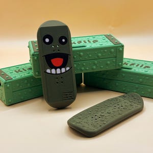 Puede incluir: Monopatines de dedo verde oliva con un diseño de cara de dibujos animados y cajas de almacenamiento verdes a juego. El monopatín tiene una cara sonriente con dientes blancos y una lengua roja. Las cajas tienen la palabra "KFlip".