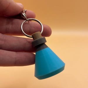May include: A blue, grey, and brown potion bottle keychain charm. The charm has a silver keyring and clasp. The bottle has a conical blue base, a grey midsection, and a brown stopper.