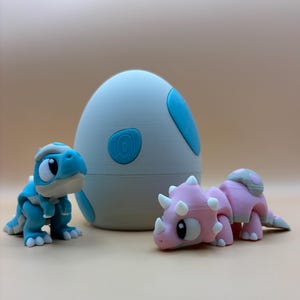 May include: Two small dinosaur toys, one blue and one pink, are sitting in front of a white egg-shaped object with blue accents. The egg-shaped object has a textured surface.
