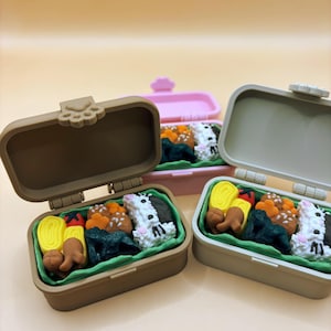 Op de afbeelding: Drie miniatuur bento-boxen, elk gevuld met kleurrijke voedingsmiddelen. De dozen zijn bruin, roze en grijs, met open deksels. Het voedsel omvat sushi, groenten en andere kleine, gedetailleerde voedselreplica's.