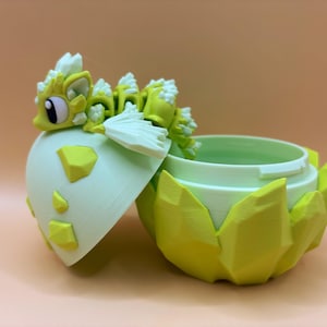 May include: A green and white 3D printed dragon figurine with a spiky back and white wings, emerging from a green and white egg-shaped container with a lid.