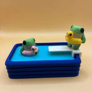 Może przedstawiać: Miniaturowa scena basenowa z dwiema zielonymi figurkami żab. Jedna żaba unosi się na różowym pływaku w kształcie flaminga, a druga stoi na białej trampolinie z żółtym pływakiem w kształcie kaczki. Basen jest niebieski z ciemnoniebieską obwódką.