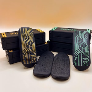 May include: A selection of black finger skateboards featuring gold and green circuit board patterns. The skateboards are displayed with small black storage boxes with yellow and green details. The word "KOKIFLIP" is visible on the boxes.