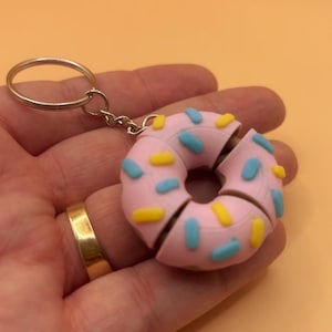 May include: A pink donut keychain with blue and yellow sprinkles. The donut is cut into four pieces and attached to a silver keyring.