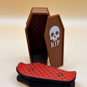 Könnte beinhalten: Eine braune Sarg-förmige Box mit offenem Deckel, mit einem weißen Totenkopf und den Buchstaben "RIP". Ein kleines, rotes Skateboard mit schwarzer Basis liegt vor dem Sarg. Der Sarg ist geöffnet und zeigt ein gemustertes Innendesign.