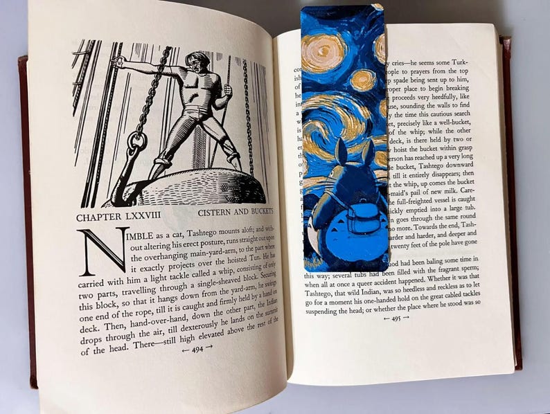 Puede incluir: Un marcap&aacute;ginas colorido con un Totoro azul sobre un cielo estrellado arremolinado, al estilo de Van Gogh. El marcap&aacute;ginas est&aacute; en un libro abierto con ilustraciones en blanco y negro.