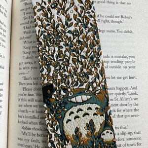 Puede incluir: Marcap&aacute;ginas artesanal con una ilustraci&oacute;n de Totoro. El marcap&aacute;ginas tiene un fondo blanco con detalles marrones y verde azulado, y est&aacute; decorado con un dise&ntilde;o de hojas y flores. Un accesorio &uacute;nico.