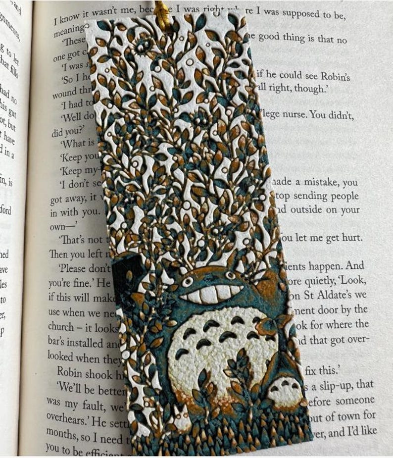 Puede incluir: Marcap&aacute;ginas artesanal con una ilustraci&oacute;n de Totoro. El marcap&aacute;ginas tiene un fondo blanco con detalles marrones y verde azulado, y est&aacute; decorado con un dise&ntilde;o de hojas y flores. Un accesorio &uacute;nico.