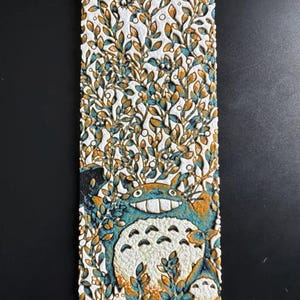 Puede incluir: Un marcap&aacute;ginas rectangular con un dise&ntilde;o texturizado de Totoro de Mi Vecino Totoro. El marcap&aacute;ginas tiene un fondo blanco con una ilustraci&oacute;n detallada de Totoro en tonos azules, naranjas y blancos, rodeado de follaje. Una cuerda dorada est&aacute; unida en la parte superior.