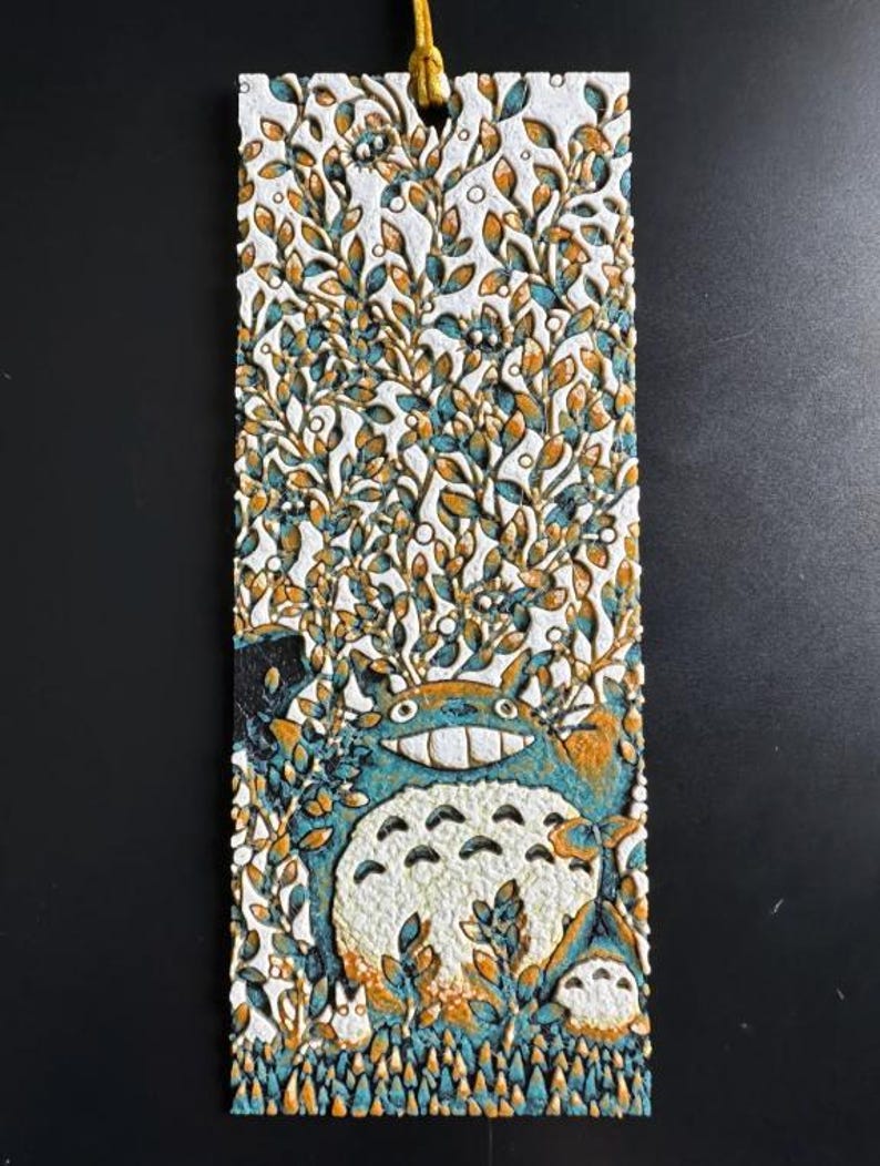 Puede incluir: Un marcap&aacute;ginas rectangular con un dise&ntilde;o texturizado de Totoro de Mi Vecino Totoro. El marcap&aacute;ginas tiene un fondo blanco con una ilustraci&oacute;n detallada de Totoro en tonos azules, naranjas y blancos, rodeado de follaje. Una cuerda dorada est&aacute; unida en la parte superior.