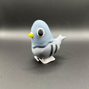 May include: A gray and white wind-up toy pigeon with black stripes. The toy has a yellow beak and large black eyes.