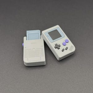 Pode incluir: Um console de jogos portátil cinza e um suporte de cartucho de jogo correspondente. O console tem uma tela, um direcional e botões roxos. O suporte do cartucho está aberto, revelando um cartucho de jogo.