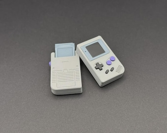 May include: A grey handheld gaming console and a matching game cartridge holder. The console has a screen, directional pad, and purple buttons. The cartridge holder is open, revealing a game cartridge.