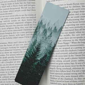 Könnte beinhalten: Rechteckiger Lesezeichen mit einem Waldmotiv. Der obere Teil zeigt einen hellblauen Himmel, der sich in einen dunkelgrünen Wald am unteren Rand verwandelt. Das Lesezeichen liegt auf gedrucktem Text.