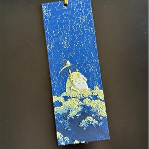 Puede incluir: Un marcap&aacute;ginas azul con una superficie texturizada, que representa una escena de Mi Vecino Totoro. El marcap&aacute;ginas presenta un dise&ntilde;o de cielo estrellado y los personajes Totoro y un Totoro m&aacute;s peque&ntilde;o bajo un paraguas, con un dise&ntilde;o de nube en la parte inferior.