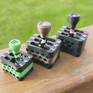 Pode incluir: Três miniaturas de manípulos de mudança de velocidades. Os manípulos são pretos com detalhes em verde, castanho e rosa. As bases das mudanças são pretas com um design em favo de mel.