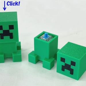 May include: Three green, cube-shaped mechanical keyboard keycaps featuring a pixelated black face design. One keycap is fully assembled, one shows the internal blue switch mechanism, and one is placed on its side with the face visible.