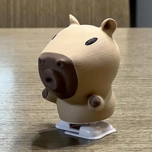May include: A tan wind-up toy capybara with brown eyes and a brown nose. The toy is on a wooden surface.