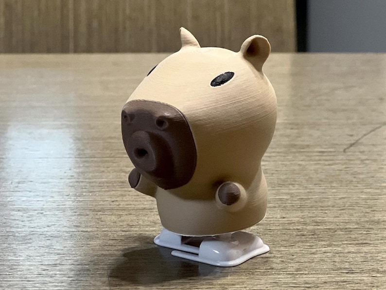 May include: A tan wind-up toy capybara with brown eyes and a brown nose. The toy is on a wooden surface.