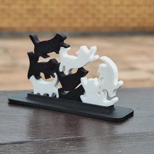 May include: A black and white cat sculpture. The sculpture features several stylized cat figures in various poses, stacked on top of each other. The base is a rectangular shape. The background is blurred.