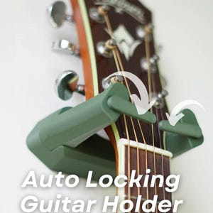 May include: Acoustic guitar headstock with a green auto-locking guitar holder. The guitar has brown wood, white accents, and metal tuning pegs. The text "Auto Locking Guitar Holder" is displayed in white.