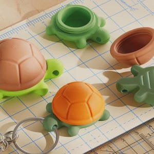 May include: A collection of turtle-shaped silicone containers and a keychain. The items feature green, orange, and brown shells. One turtle is attached to a metal keyring. A ruler with centimetre markings is visible, showing the scale of the objects.