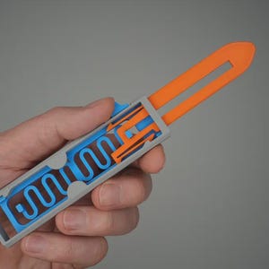 Könnte beinhalten: Ein 3D-gedrucktes, abstraktes Objekt, das in einer Hand gehalten wird. Das Objekt ist hauptsächlich grau, mit orangefarbenen und blauen Innenteilen. Der orangefarbene Abschnitt erstreckt sich aus dem grauen Rahmen, der blaue Teil hat ein wellenförmiges Design.