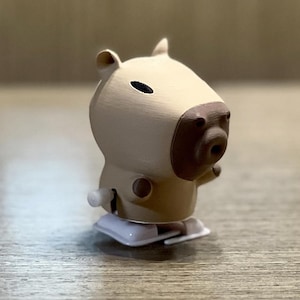 May include: A beige and brown wind-up toy capybara with a black eye. The toy has a white base and a key to wind it up.