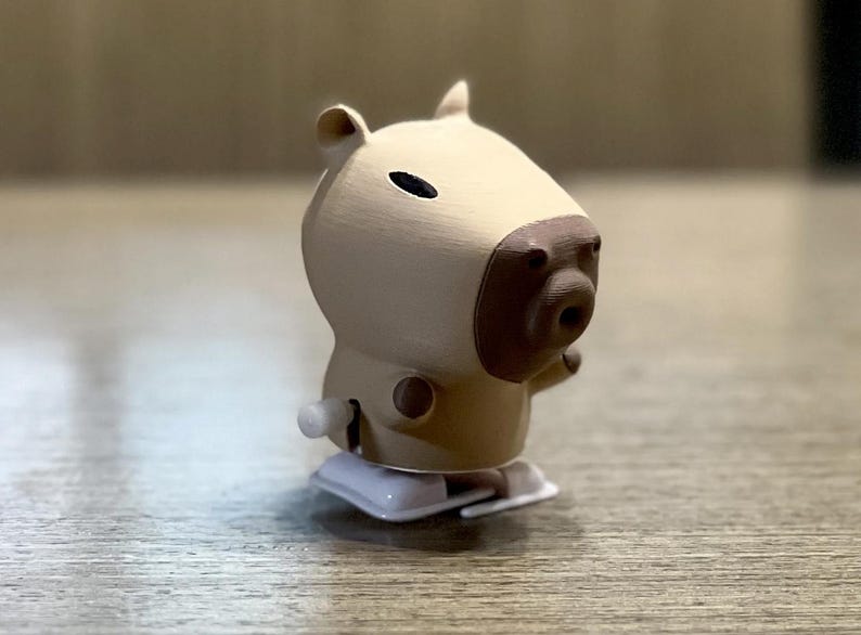 May include: A beige and brown wind-up toy capybara with a black eye. The toy has a white base and a key to wind it up.
