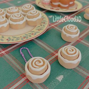 Könnte beinhalten: Mehrere Miniatur-Zimtschnecken mit weißem Zuckerguss sind auf einem Teller angeordnet und auf einer grün karierten Oberfläche verstreut. Eine Rolle ist an einer lila Perlenkette befestigt. Der Text "LittleCloud3D" ist sichtbar.