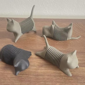 May include: Four cat-shaped figurines with ribbed, accordion-style bodies. Three of the figures are light gray, and one is dark charcoal gray. Each cat has a pointed tail, ears, and a smooth head, with a flexible, segmented torso design.