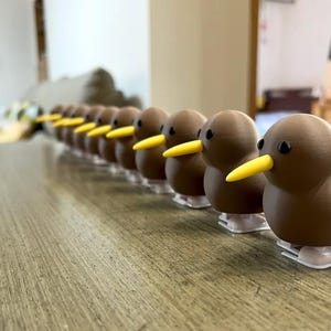 May include: A line of brown and yellow toy kiwis lined up on a wooden surface. The kiwis have yellow beaks and black eyes.