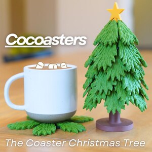 May include: A green Christmas tree shaped coaster with a star on top. The coaster is made of a soft, flexible material and is designed to hold a mug or cup. The text "Cocoasters" is visible in the background. The coaster is sitting on a wooden surface.