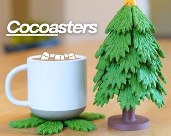 May include: A green Christmas tree shaped coaster with a star on top. The coaster is made of a soft, flexible material and is designed to hold a mug or cup. The text "Cocoasters" is visible in the background. The coaster is sitting on a wooden surface.