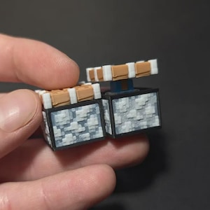 May include: Two small, square, block-shaped items featuring a pixelated gray and white stone texture on the sides. Each block is topped with a tan and white rectangular lid. The items have a black frame and a small blue accent visible on one side.