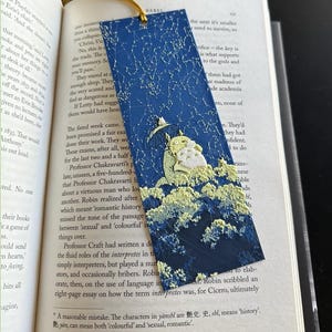 Puede incluir: Marcap&aacute;ginas azul marino con borla dorada. El marcap&aacute;ginas presenta una ilustraci&oacute;n caprichosa de un personaje con paraguas, sobre un dise&ntilde;o de cielo estrellado y nubes. El marcap&aacute;ginas est&aacute; colocado en un libro abierto.