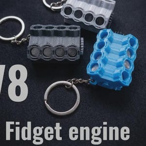 Może przedstawiać: Breloki z modelami silników V8 w kolorze szarym i niebieskim, ze srebrnymi kółkami na klucze. Tekst "V8 Fidget engine" jest wyświetlany na biało. Te breloki z silnikiem drukowane w 3D to wyjątkowy dodatek.