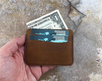 May include: A brown leather cardholder with two American Express cards and a one-dollar bill peeking out. The cardholder is rectangular with stitched edges and a minimalist design, perfect for carrying essential cards and cash.