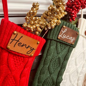 May include: Three knitted Christmas stockings with personalized leather name tags. The stockings are red, green, and white with a cable knit pattern. The names on the tags are Henry, Lucas, and a third name that is not visible.