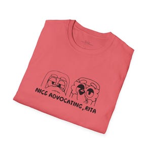 May include: A coral-colored t-shirt featuring a black graphic of two cartoon characters with expressive eyes. The text "NICE ADVOCATING, RITA" is printed below the characters. The shirt is made of soft material.
