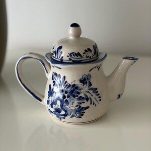 Może przedstawiać: Kremowy ceramiczny czajniczek z ręcznie malowanymi niebieskimi wzorami kwiatowymi. Czajniczek posiada pasującą pokrywkę z małym uchwytem, zakrzywioną rączkę oraz zwężający się dzióbek. Niebieskie wzory składają się ze szczegółowych liści i płatków.