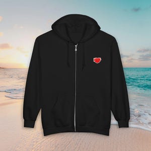 May include: Black zip-up hoodie with a hood and front pockets. A small, embroidered red heart with a smiling face is on the upper left chest. The hoodie is displayed on a beach with a sunset in the background.